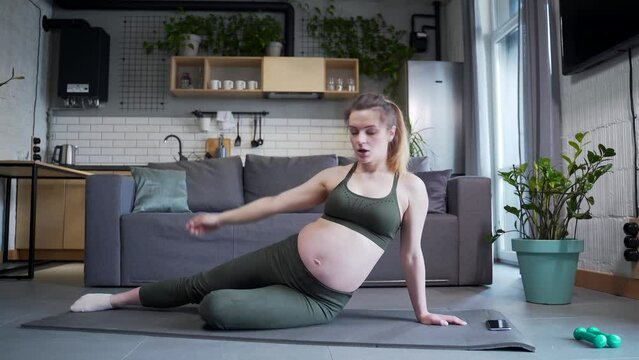 A young pregnant woman is doing gymnastics or pilates exercises. practice yoga stretching exercises during pregnancy. Sitting on a mat in the living room at home. Indoor fitness and workout