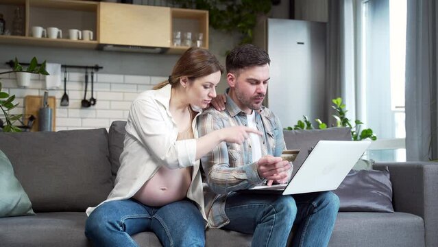 Expectant married couple pregnant wife and husband shopping online paying bills using laptop computer and credit card sitting at home on sofa. Family during pregnancy, buying goods in an online store
