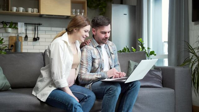 young married couple, pregnant wife and husband use laptop while shopping online sitting at home on sofa in room Choosing goods for newborn child on Internet Family Browsing computer during pregnancy