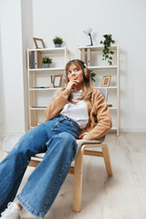 Fototapeta premium Dreaming happy young blonde lady in warm sweater in headphones looks aside listen fav songs sitting in armchair at modern home interior. Music time Relaxing Cool playlist Concept. Copy space