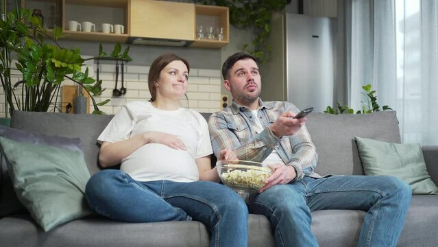 Bored couple at home choose a television show to watch and switch channels with a TV remote control. Young family husband and pregnant wife cannot choose a show. watching news or film during pregnancy