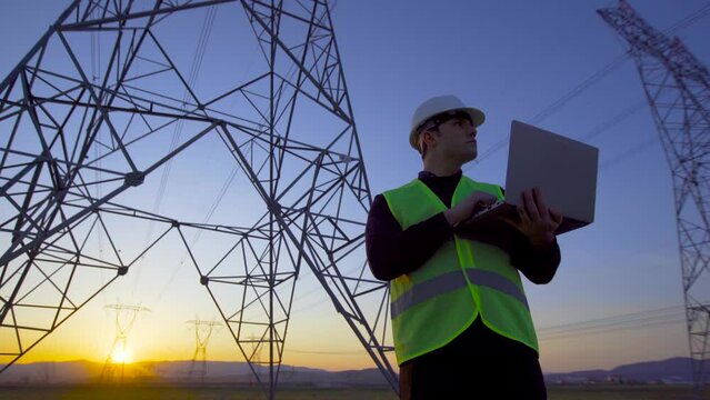 Electrical engineer working in front of high voltage lines.
Engineer working on laptop at sunset and looking at electric poles.

