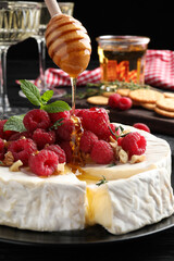 Pouring honey onto brie cheese served with raspberries and walnuts on black plate, closeup
