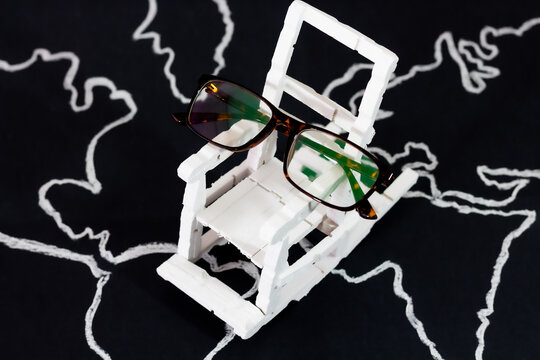 Concept World Day of Older Persons. Reading glasses on an old miniature rocking chair in white on a world map drawn with chalk on a black background.