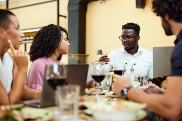 A multicultural group of businesspeople is sitting in a restaurant during business lunch and listening to a manager who explains issues about the project.