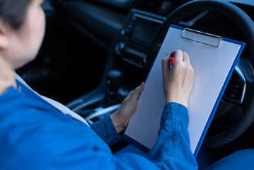 Close-up of an unrecognizable mechanic in a blue jumpsuit uniform, sitting inside the car behind the steering wheel, holding a checklist clipboard, evaluating. Car care, physical maintenance concept.