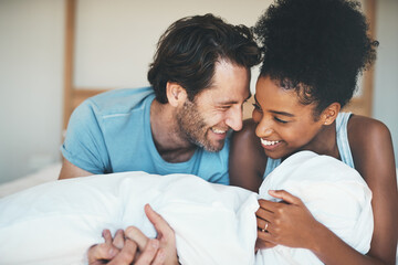 Happy, loving and funny interracial couple smiling and laughing while lying in bed and sharing a...