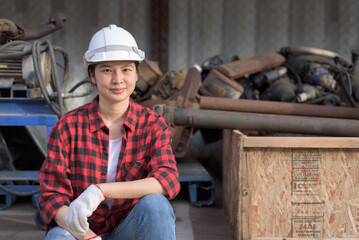 Portrait with copy space, Asian woman wearing red lumberjack shirt and safety hardhat sitting, smiling, relaxing, looking at camera. Female worker or engineer portrait at garage or workshop.