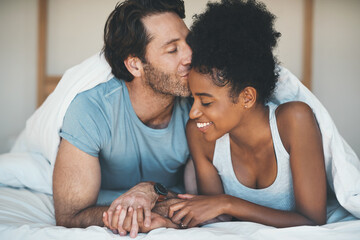 Happy, carefree and loving couple share a kiss lying in bed together in the morning. Interracial,...