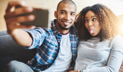 Funny and silly couple taking selfie and making goofy face at phone for social media post while relaxing. Boyfriend and girlfriend having fun and enjoying time together at home and sharing it online - obrazy, fototapety, plakaty
