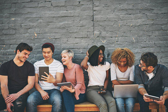 Group of friends on digital devices and social media sitting together while smiling and talking. Technology obsessed online remote workers taking a break. Happy casual creative team outside