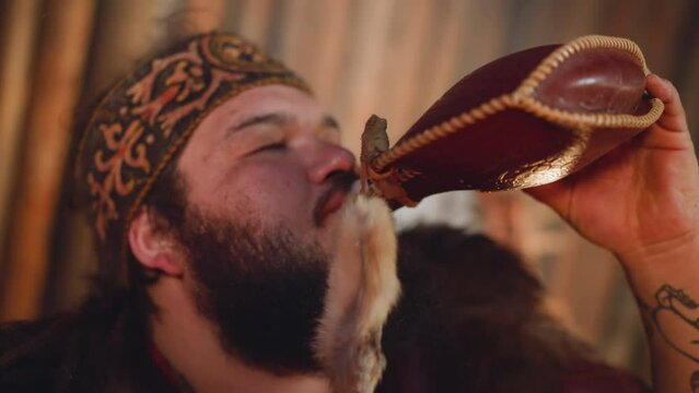 Tired plump man in national Altai clothes drinks from leather waterskin in cabin closeup. Traditional tribal pack for liquids carriage. Ethnic festival