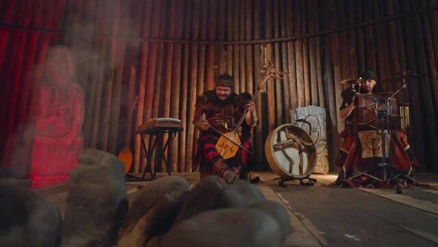 Folk rock band plays traditional Altai music with ancient instruments near stone statue in ail house. Emotional integration of old culture in modern life