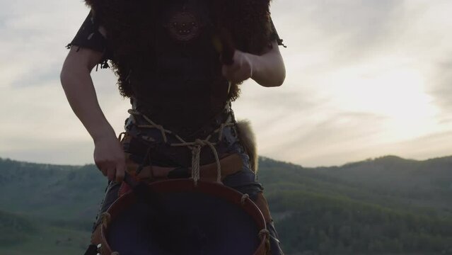 Drummer in leather vest with fur taps tambourine against distant mountains at sunset closeup. Man performs Altai music at ethnic festival