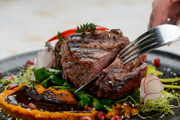 meat steak grilled on a black plate with salad is cut with utensils in female hands

