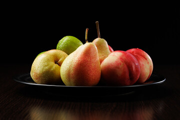 Ripe fruits in a plate close-up on a black background. Side view, selective focus. Pears, nectarines and limes.