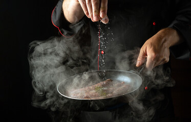 The chef adds salt to a hot frying pan with sausages. Menu or recipe concept for hotel on black background