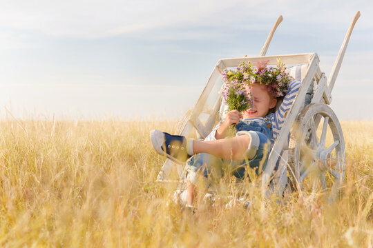 Child plays in a field on a summer day. Little girl with a bouquet of flowers in a garden wheelbarrow against the blue sky. Cheerful and happy kid dreaming on the grass.