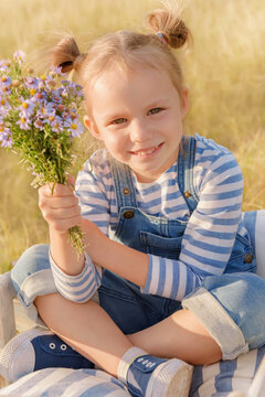 Child plays in a field on a summer day. Little girl with a bouquet of flowers in a garden wheelbarrow against the blue sky. Cheerful and happy kid dreaming on the grass.
