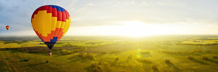 Colorful hot air balloons fly over the fields at sunset on a warm autumn evening. Panorama of a beautiful landscape in the sunlight from a bird's eye view.