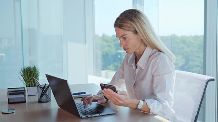 An office worker makes a calculation, a financial report, uses wireless Internet technologies, a phone and laptop. Businesswoman sit in office hold smartphone enters data into the computer pc indoor.