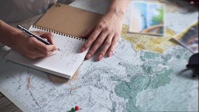 A table with a map on it. A woman makes a travel itinerary. Traveler with travel items: sunglasses and landscape photos. Top view of a tourist planning and writing down a route in a notebook.
