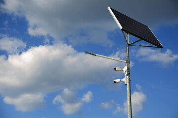 Słup z dwoma kamerami, panel słoneczny i lampa uliczna na tle nieba.
