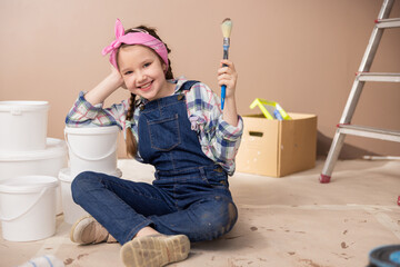 Joyful child in denim clothes soiled with brown wall paint sits on the floor holding a paintbrush...