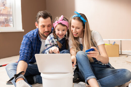 Happy family sits on floor of purchased apartment they just renovated browsing room inspirations on their laptop choosing paint colors for walls buying decorations for room paying with debit bank card