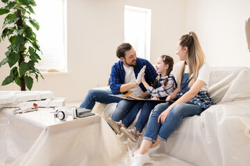 Young family sitting on a sofa in a room undergoing renovation. Father and daughter give each other high fives. Everyone is happy smiling because they finally chose new wall color for living room.