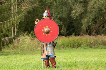 Knight soldier in armour holding red wooden shield during medieval re-enactment in front of green...