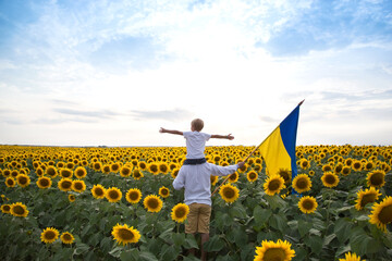 dad and son, who sits on his shoulders with arms apart, stand with flag of Ukraine on blooming...
