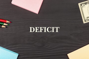 DEFICIT - text, money dollars, stickers and colored pencils on a black wooden table. Business concept: buying, selling, commerce (copy space).