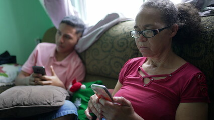 Two hispanic family members sitting on couch looking at cellphones. A senior woman looking at phone...