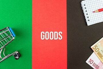 GOODS - word (text) and euro money on a table of different colors, a trolley, a basket of grocery notepad and a red pencil. Business concept, buying, selling, supermarket, store (copy space).