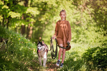 Siberian husky and his little girl owner walk together along the road in the summer park.