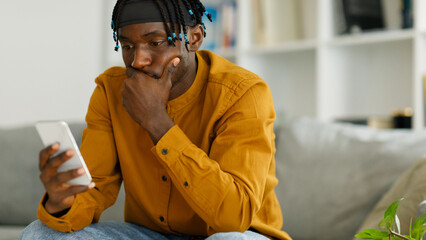 Sad young black american guy sitting on the couch, looking at the phone,