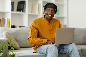 Happy smiling african american man using laptop internet, posing, looking at camera while sitting on sofa at home. , free copy space, mockup