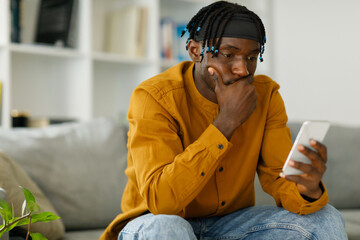 Upset black african man reading bad news in message looking at phone screen sitting on couch
