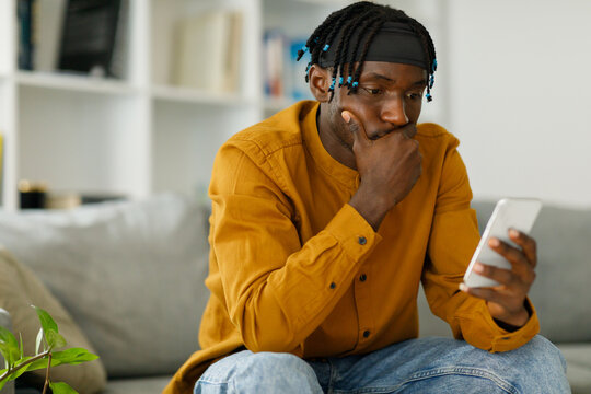Upset black african man reading bad news in message looking at phone screen sitting on couch