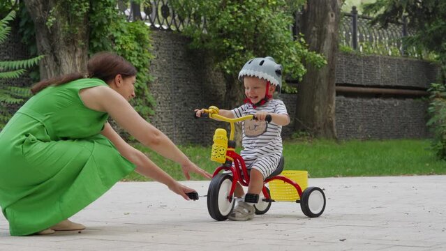 Mother teaching little child to ride tricycle for kids outdoors, toddler boy in funny bike helmet having fun playing in summer park. Outdoor playground riding activity for children.