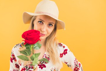 Caucasian blonde woman in sun hat and floral blouse holding up rose and looking at camera. Studio shot. High quality photo
