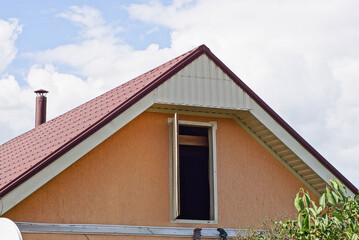 one brown attic of a private house with an open door under a red tiled roof on the street against the background of a blue sky and white clouds