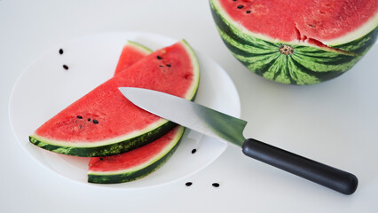 Close-up slices of fresh ripe red watermelon on a plate with a knife on the table on a white background, half of a watermelon. Ripe juicy summer berries, melon season. Macrophoto