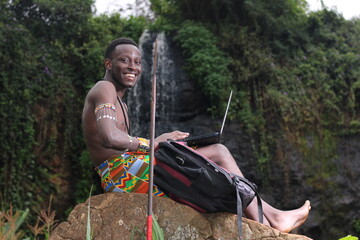 Education in Africa,online classes in Africa: A young African Masai Moran warrior sits on a rock beside a waterfall studying using a laptop 
