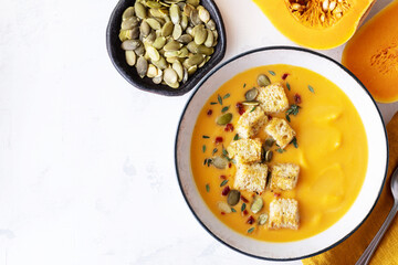 Autumn pumpkin cream soup in bowl served with seeds and crouton on white stone table top view.