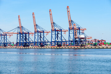 Tall gantry cranes on a commercial dock with stacks of colourful containers in background on a...