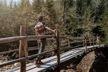 Rear on man traveler with backpack standing on bridge over river in forest. Traveling concept.