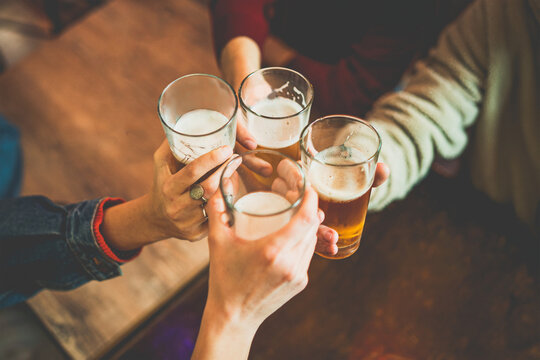 Young people celebrating with beers toasting at the irish pub - closeup crop shot of hands holding beer glasses clinking over the pub's table - friendship and alcohol lifestyle concept