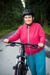 Obraz premium Elderly woman in helmet stands next to bicycle on forest road. Senior lady doing cycling in wooded place. Healthy, sportive, active lifestyle in old age